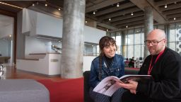 A man with glasses and a Swinburne lanyard having a conversation with a young woman in a denim jacket, both seated in a modern, open space, with the man gesturing towards a Swinburne course guide booklet.