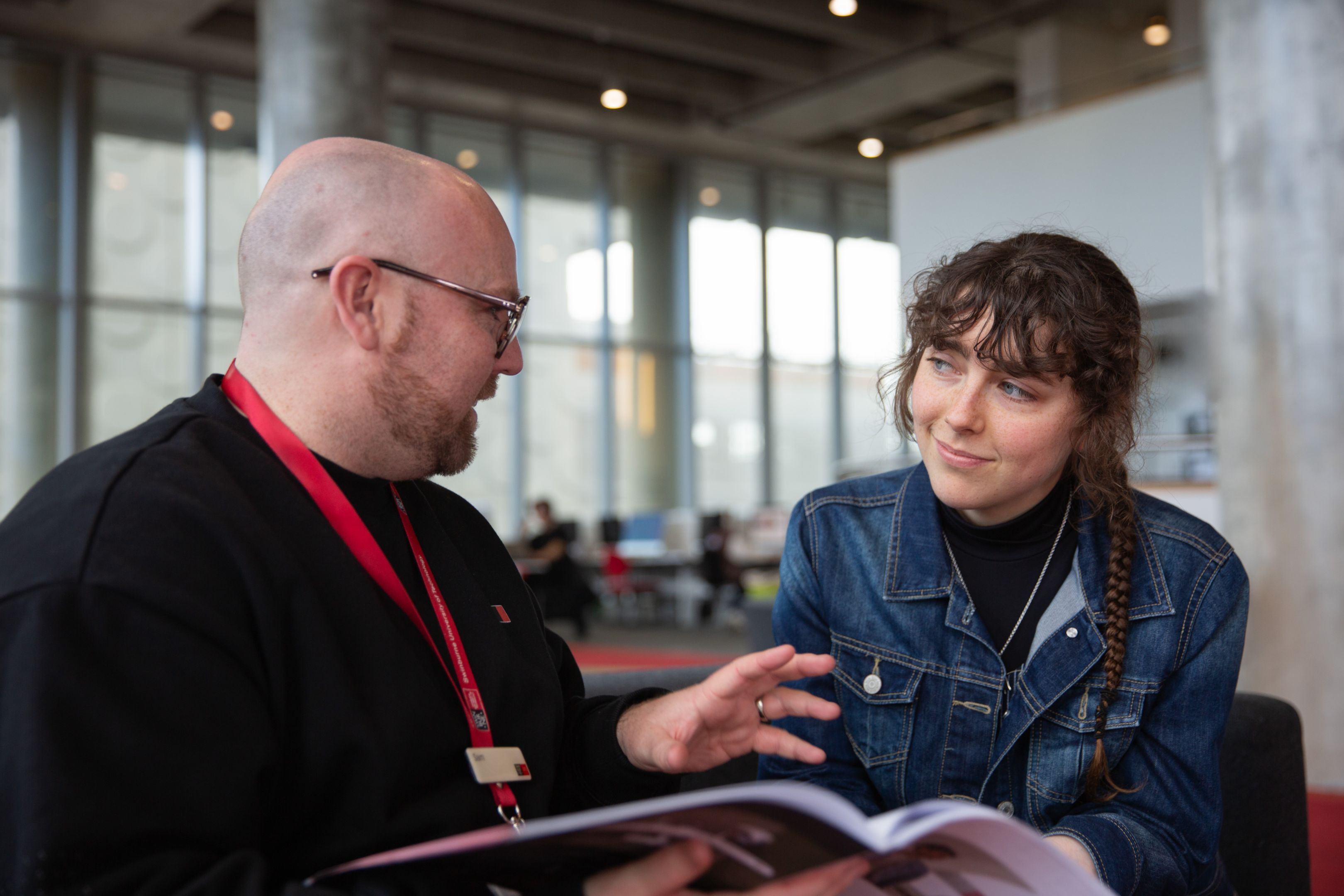 A man with glasses and a Swinburne lanyard having a conversation with a young woman in a denim jacket, both seated in a modern, open space, with the man gesturing towards a Swinburne course guide booklet.