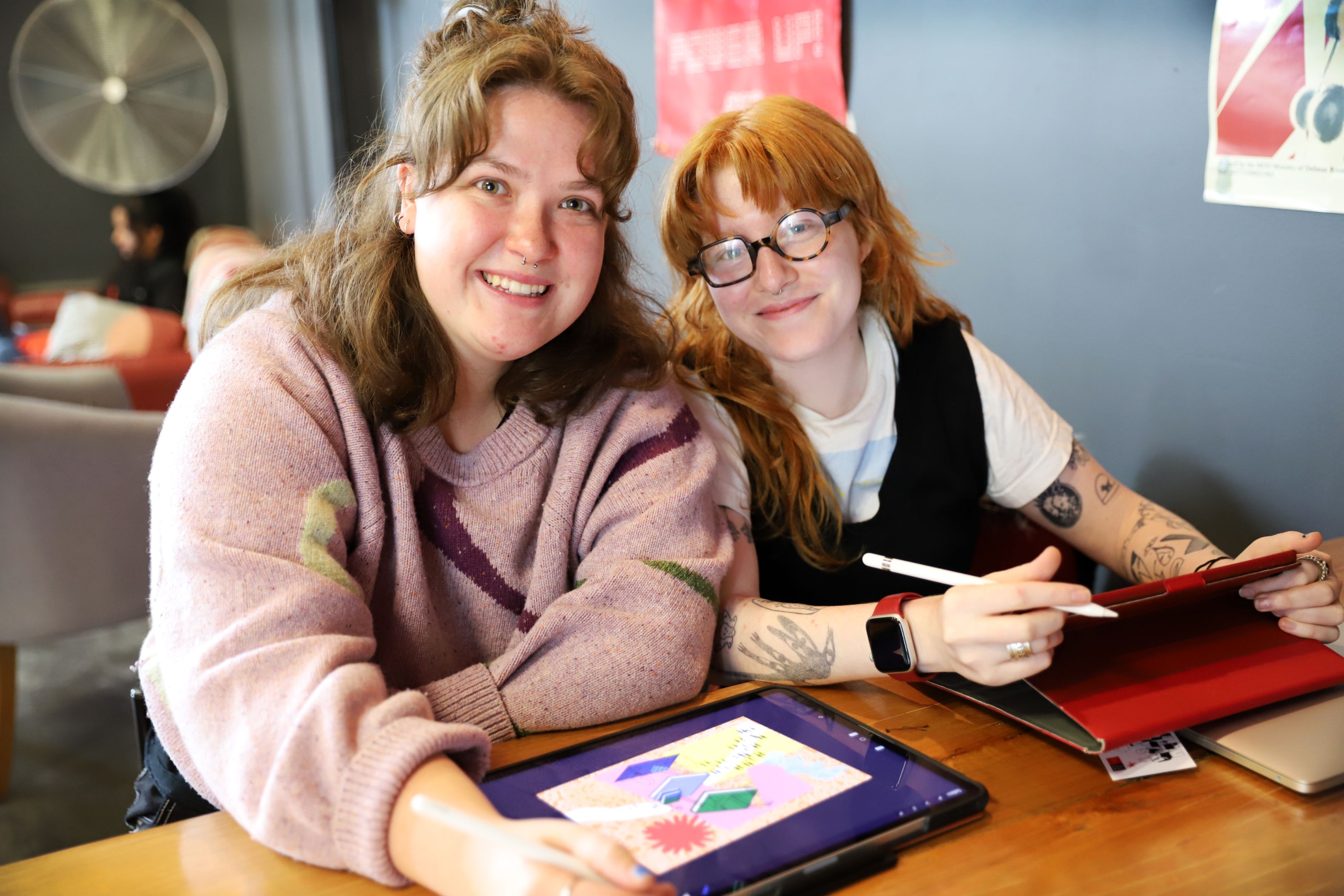 Two students smile for the camera as they work on designs on their tablets