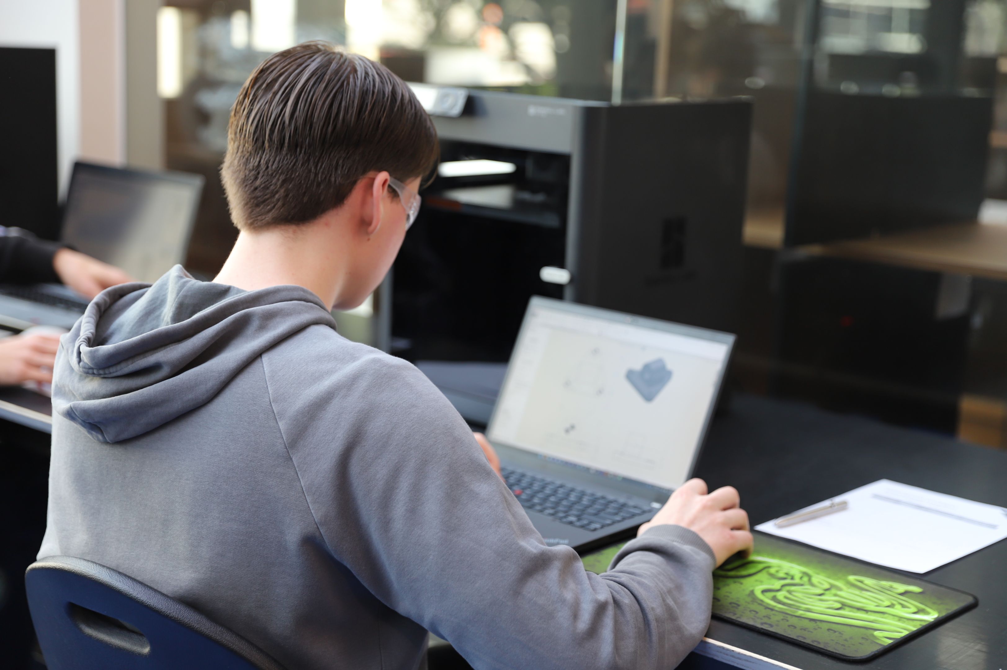 WorldSkills Regional Competition 2025 -1 Young male student sitting at a laptop at a desk, their body faced away from the camera, wearing goggles and with their hand on a mouse.
