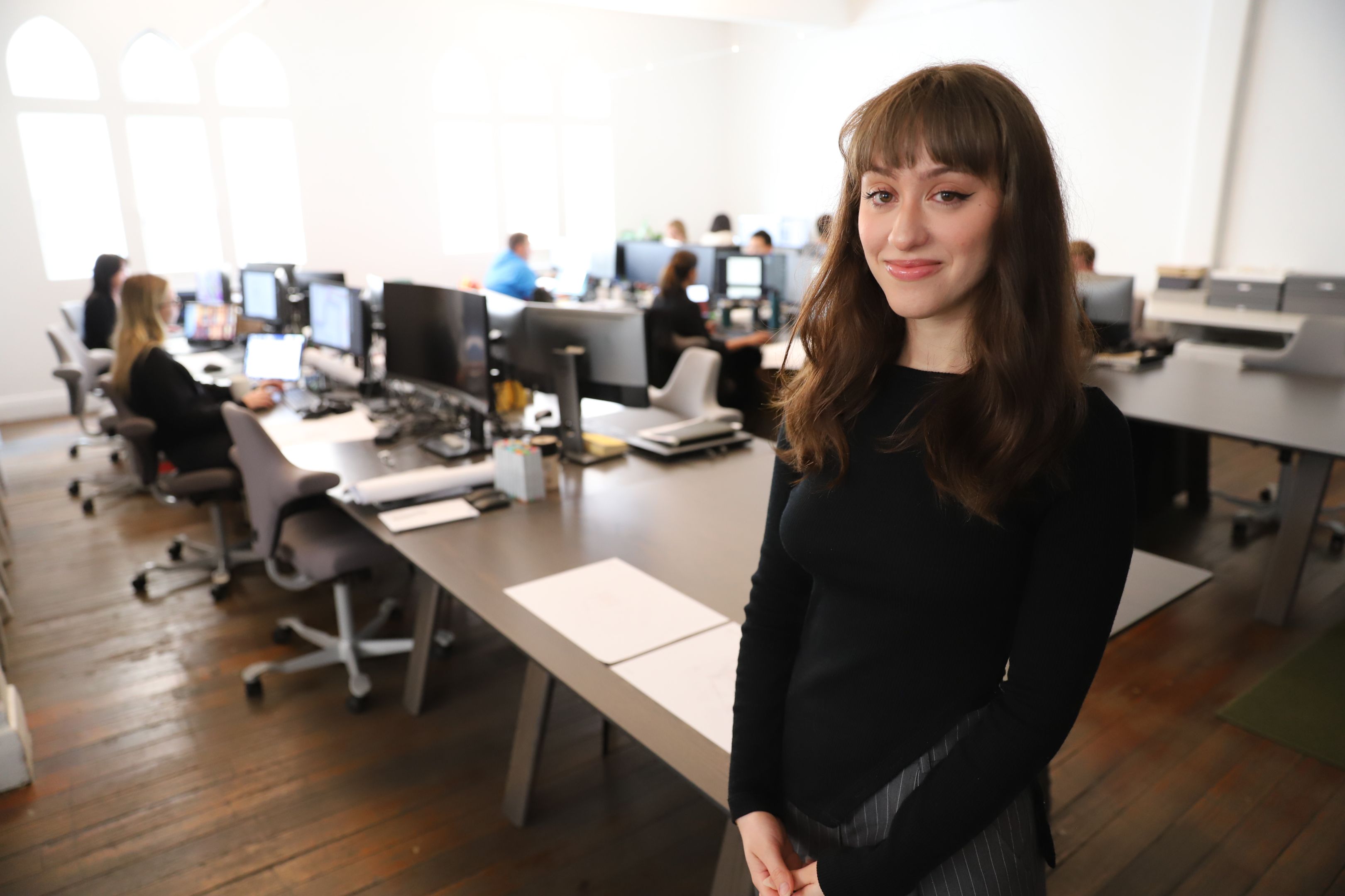 A student smiles at the camera. She is in an office, standing in front of rows of desks where people work at computers.