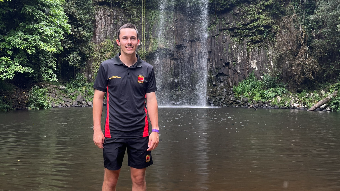 A teenager in a black Clontarf Academy shirt and shorts stands in front of a waterfall