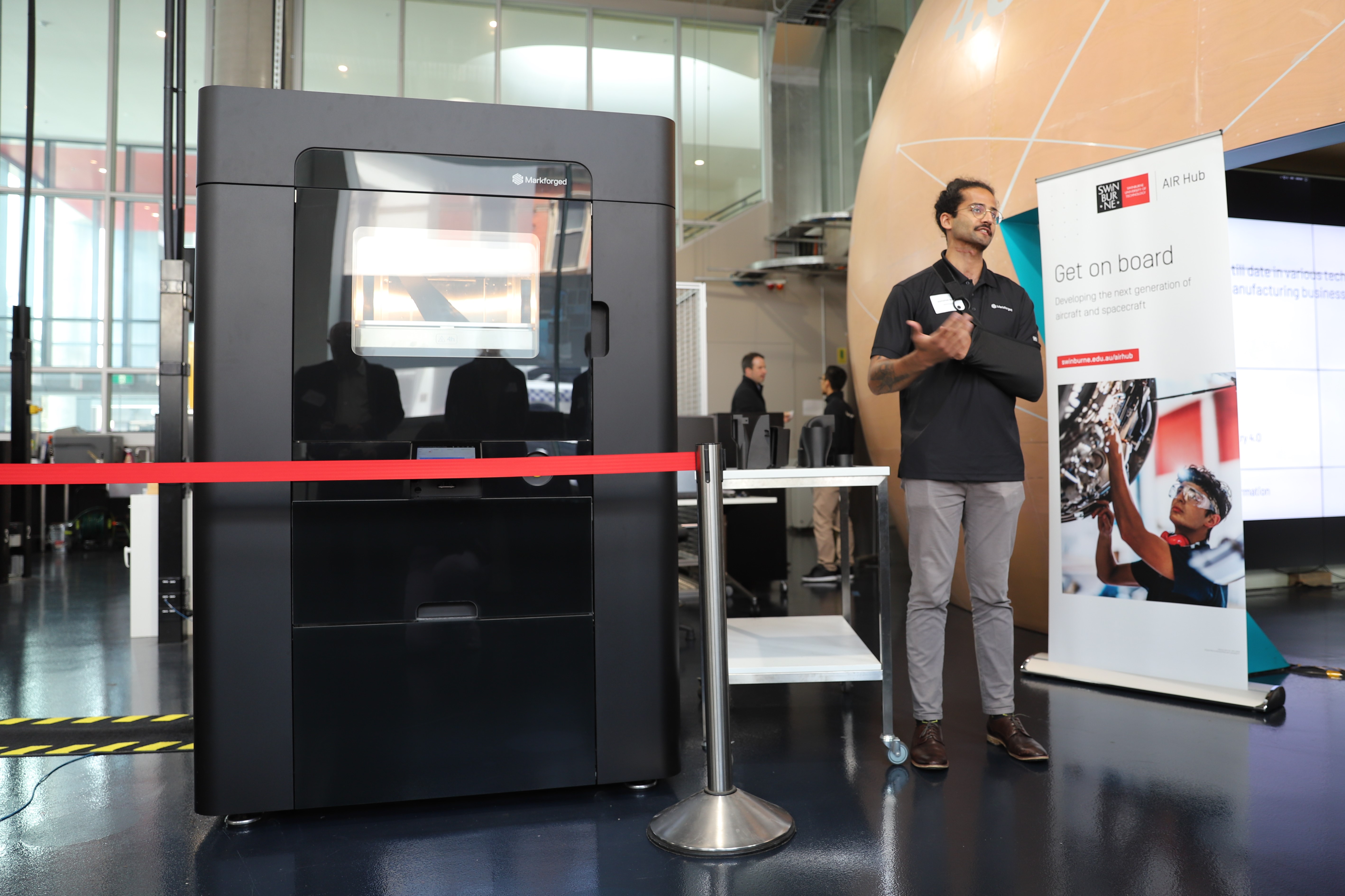 A large black 3D printer stands beside a man presenting