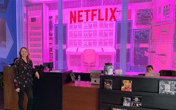 Sue wears a black shirt and pants and stands in front of the Netflix reception areas. A huge desk and chair installation can be seen behind a glass wall and the space is illuminated by pink neon light. 