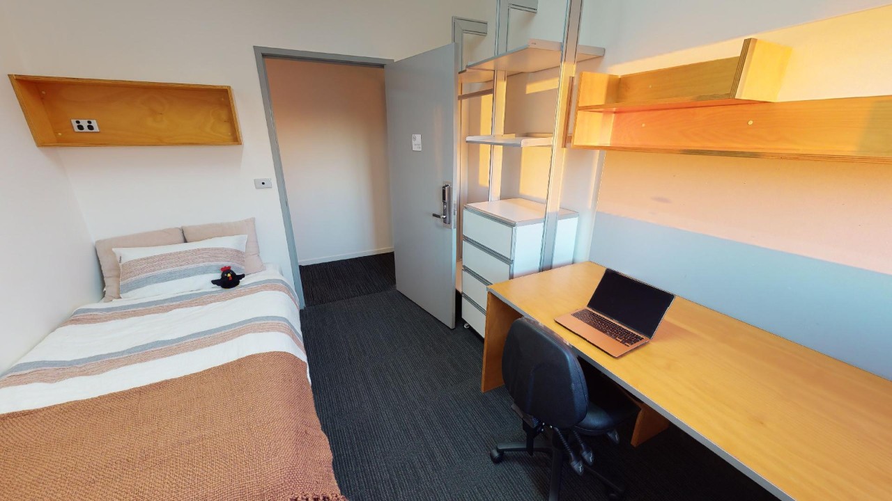 A bedroom with a bed and stripy doona cover, desk, laptop, chair and shelving