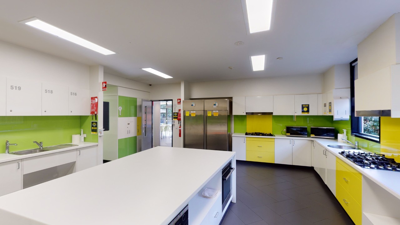 A spacious kitchen with island bench, yellow and white cupboards., green splashback and a fridge