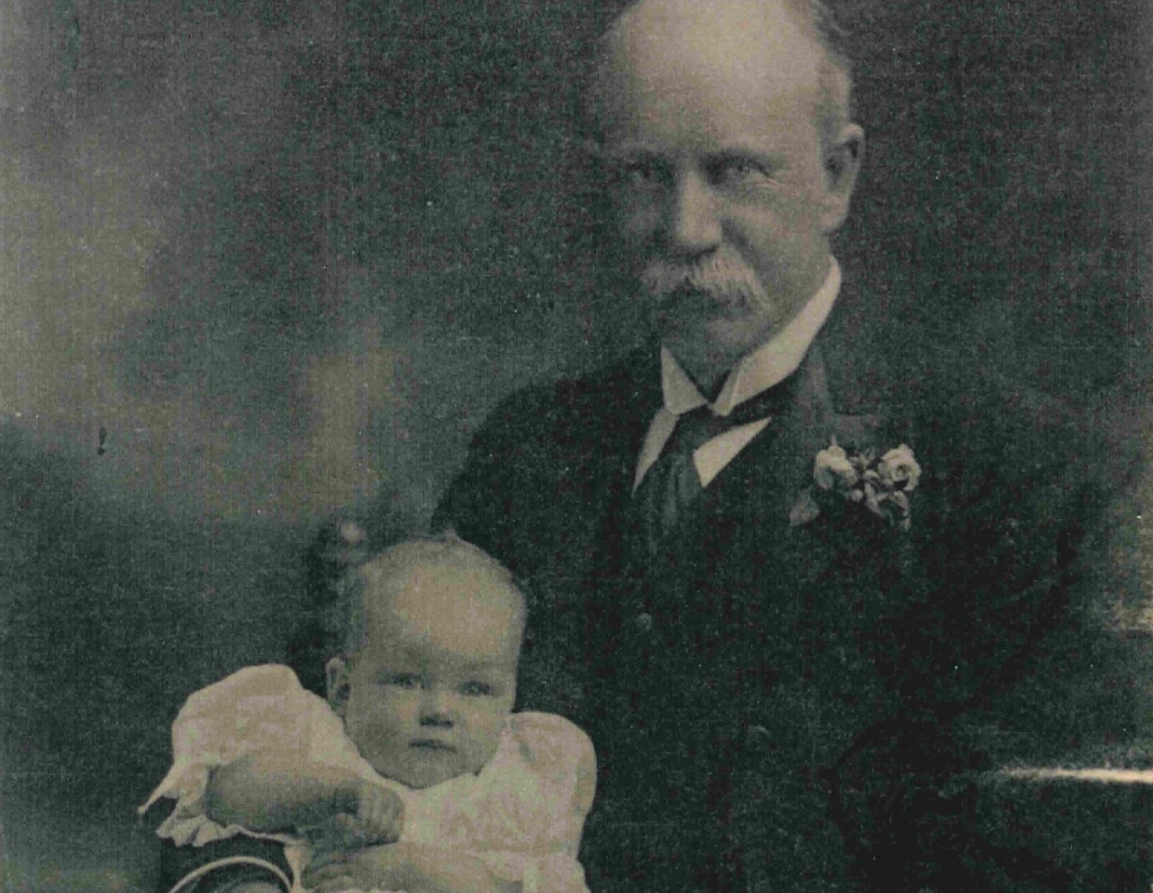 A photograph from the early 1900s of the Honourable George Swinburne and his baby daughter, Margaret who appears around six months old. She sits on his lap in a white frilly dress, he has an arm around her. 