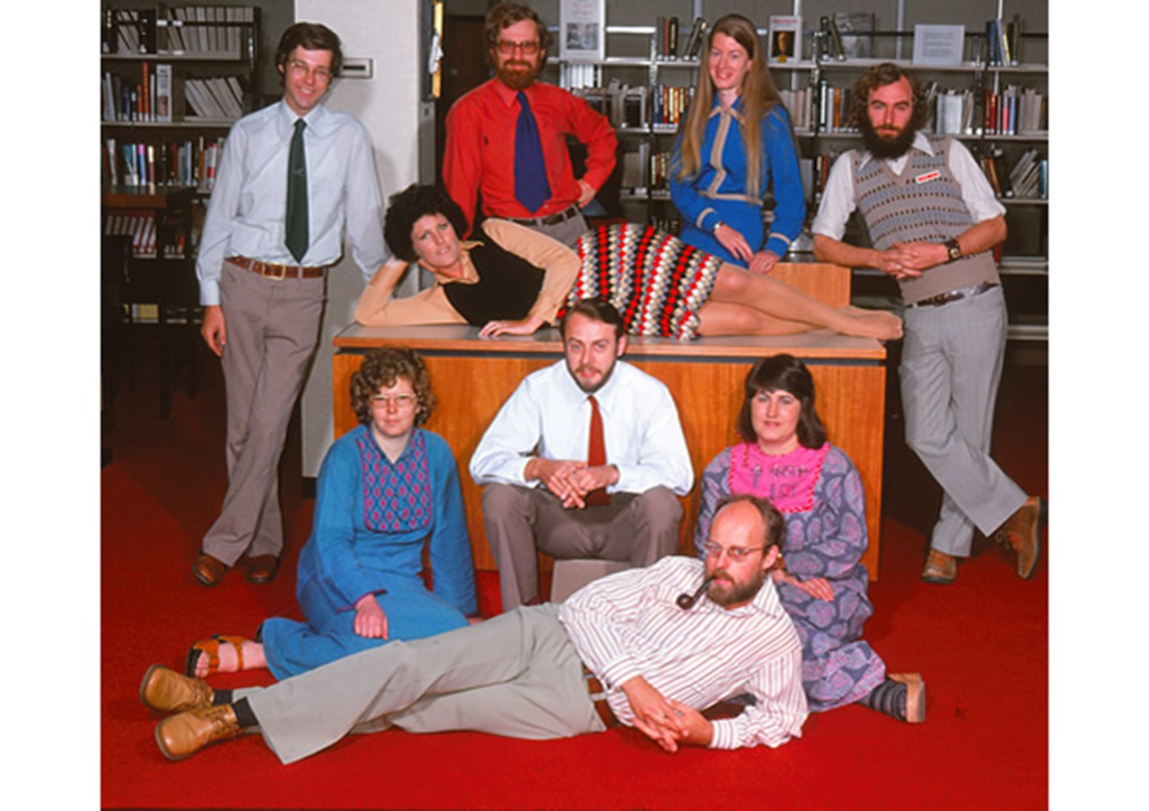 Nine librarians pose for a group photograph at the Swinburne library in 1974. They wear clothes typical of the 1970s – paisley, clogs and crochet. One lies across a desk, another lies at the front smoking a pipe! 