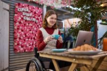 A young, smiling woman in a wheelchair holds a cup of tea while looking at something on her laptop in an outdoor garden.