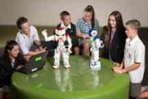 Three girls and three boys in school uniforms sitting around a green table playing with two robots