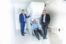Two men in suits standing next to a large neuroimaging type machine with someone sitting in the seat