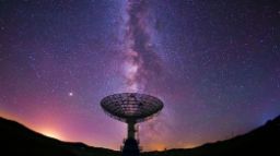The silouhette of a telescope against a night sky filled with bright stars, the milky way with colours of purple, blue, orange and pink present