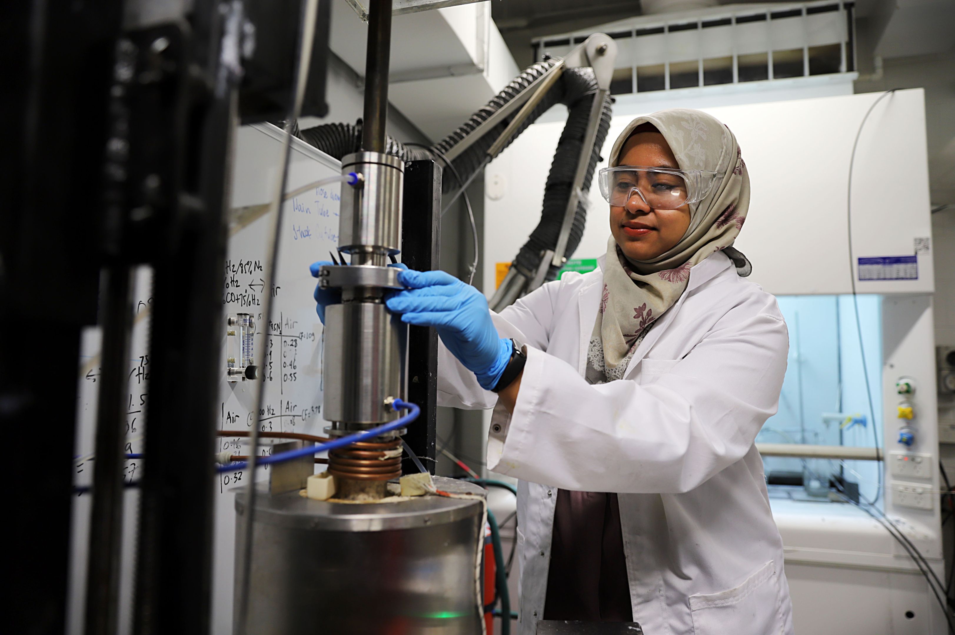 A woman in a lab coat, protective eye glasses and latex gloves operates machinery in a scientific research lab