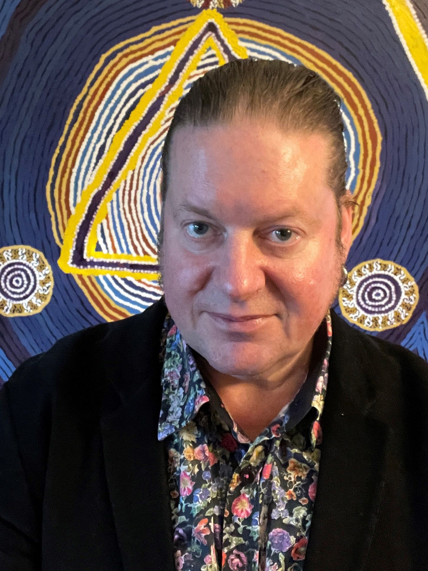 Man in floral shirt and black jacket standing in front of indigenous background