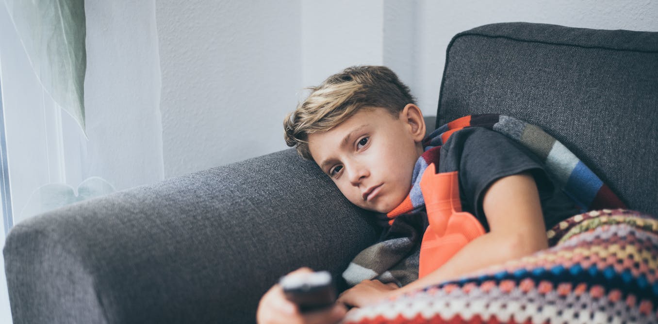 A kid wrapped in a blanket and hugging a hot bottle is holding a TV remote whilst on the couch