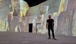 Man standing in thr Lume in front of the wall/screens projecting an artwork.