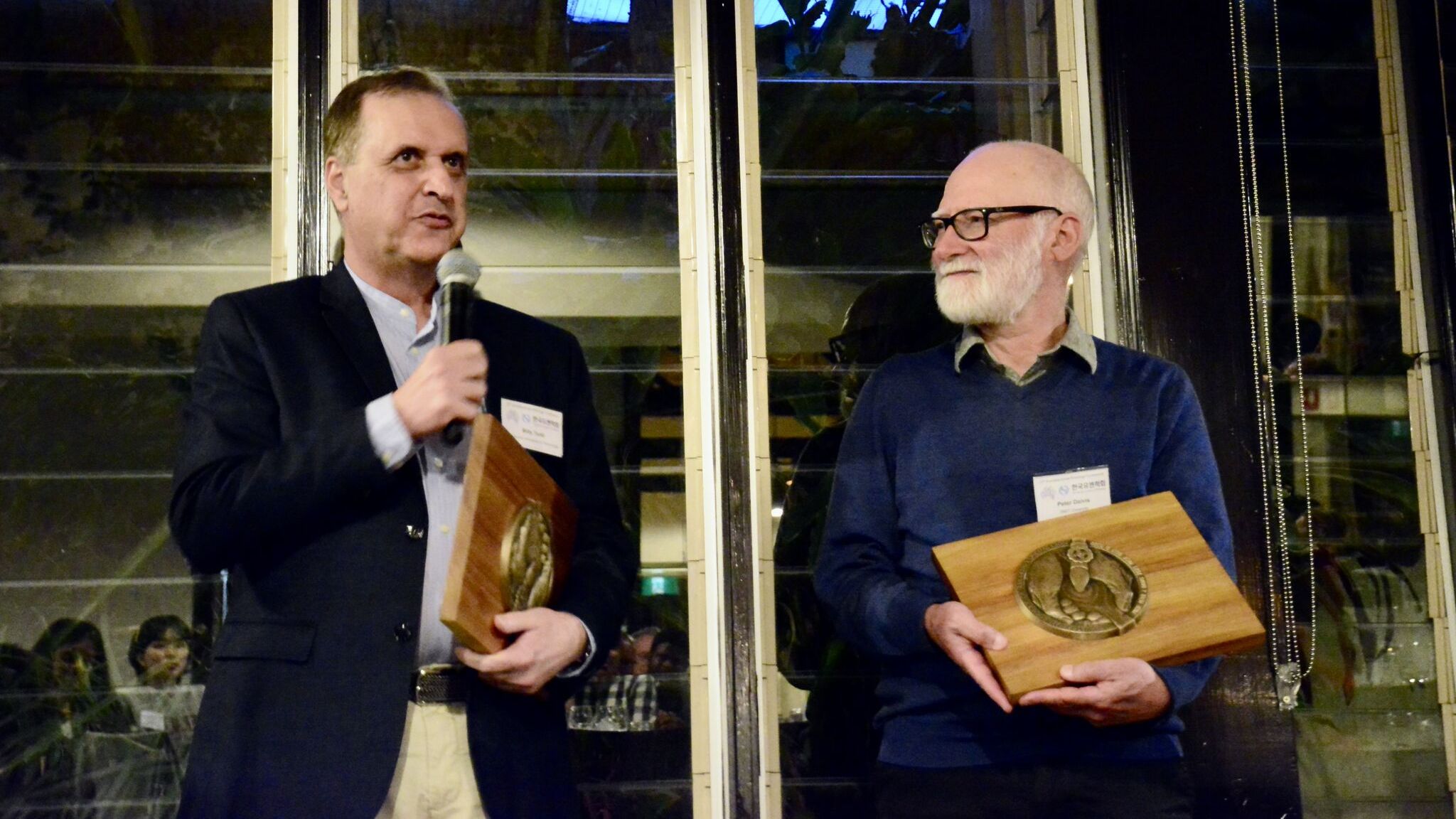 Two middle aged men in professional dress holding wooden awards, man on left is holding a microphone and speaking.