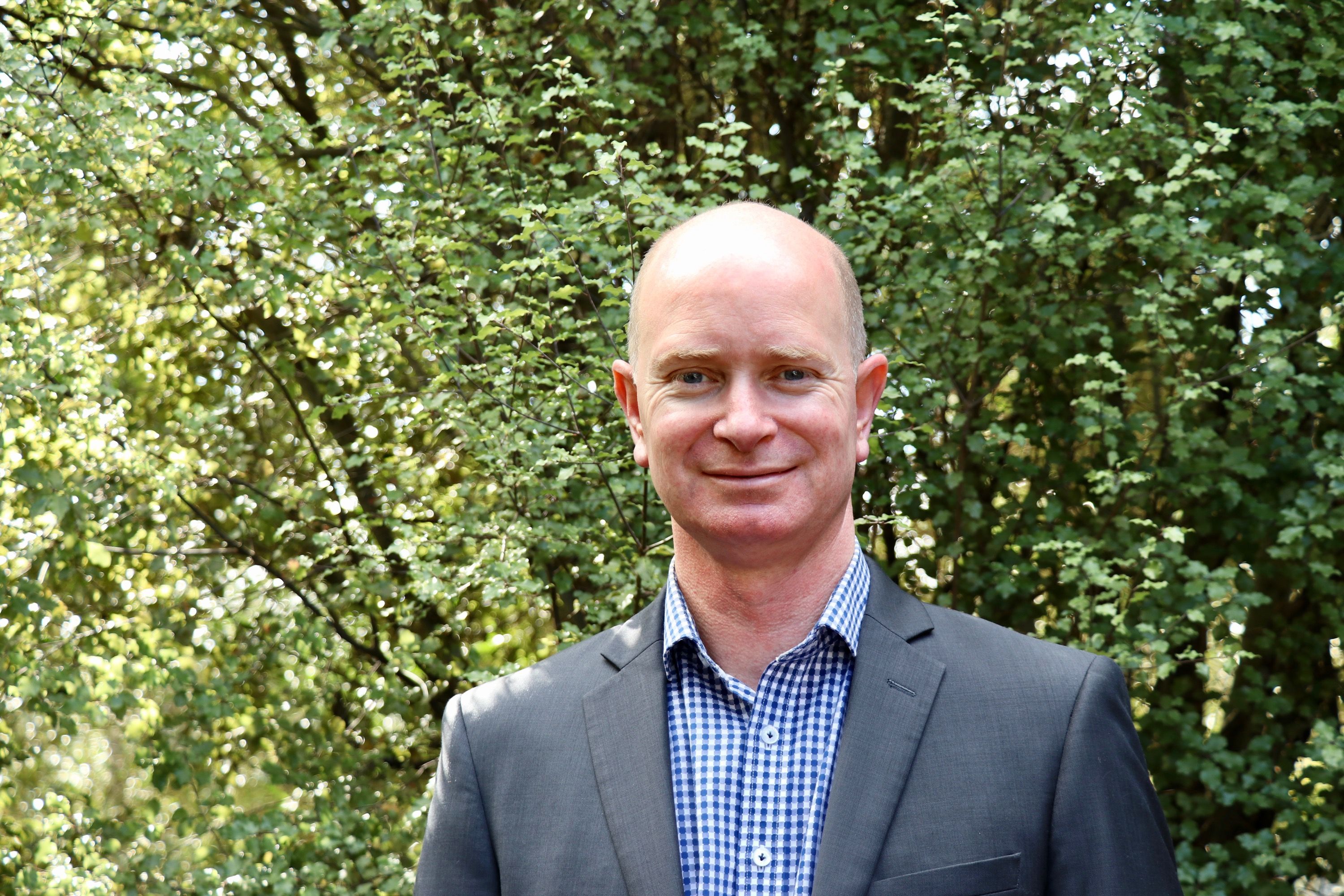 Shot of Justin Potter, middle aged man of Caucasian decent, from the chest up wearing a grey suit and blue check shirt. Leafy trees in the background.