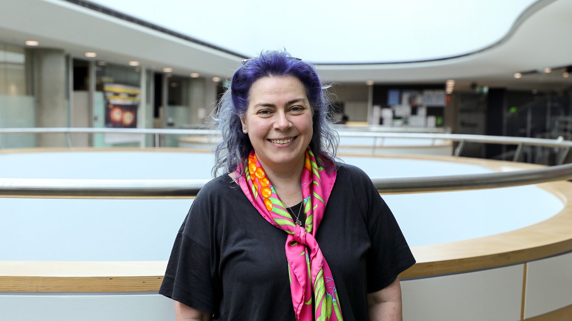 Woman with purple hair, in a black top and pink scarf smiling for the camera.