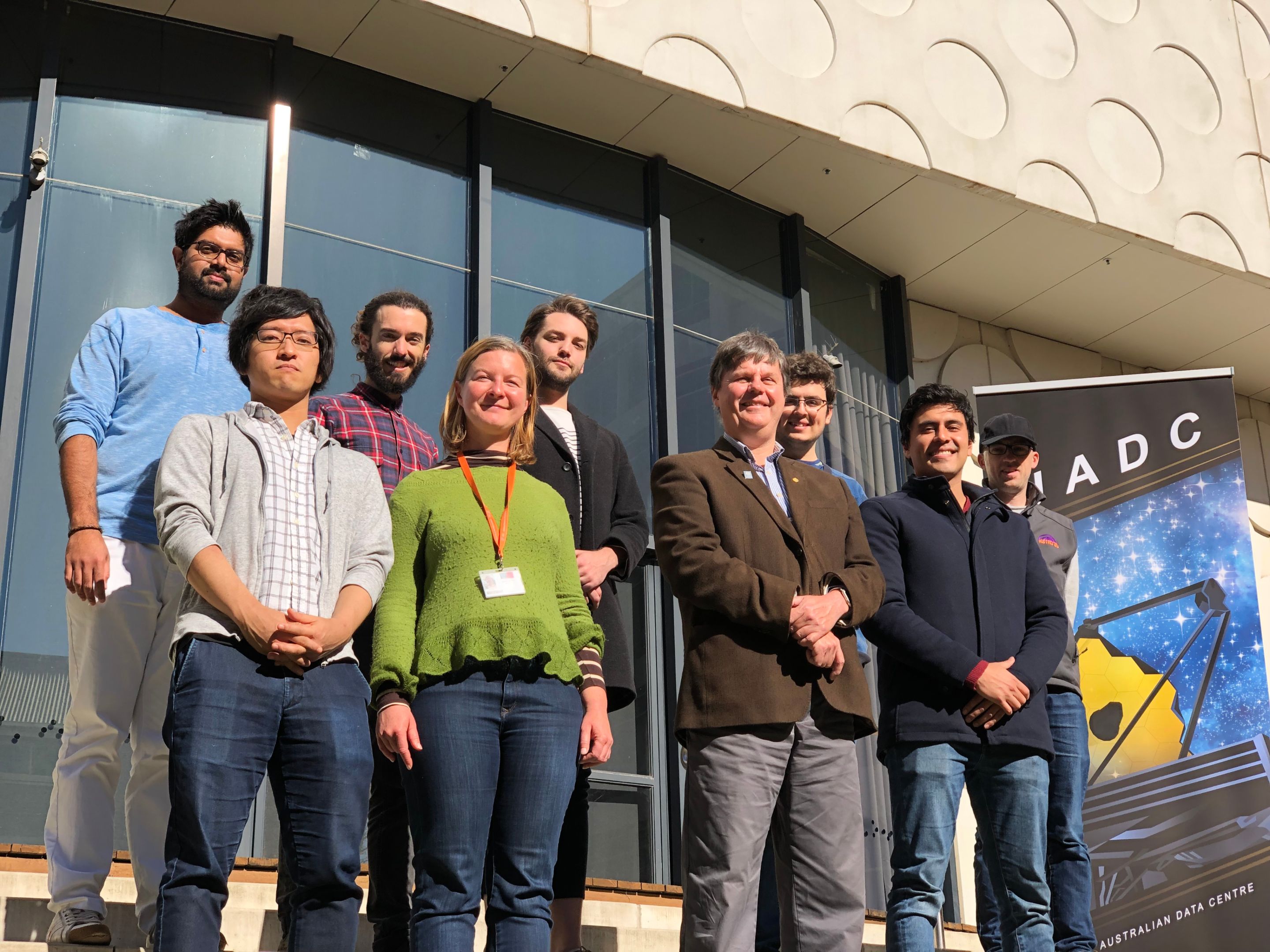 Eight men and one women from the JWST Laureate science team standing in front of glass windows