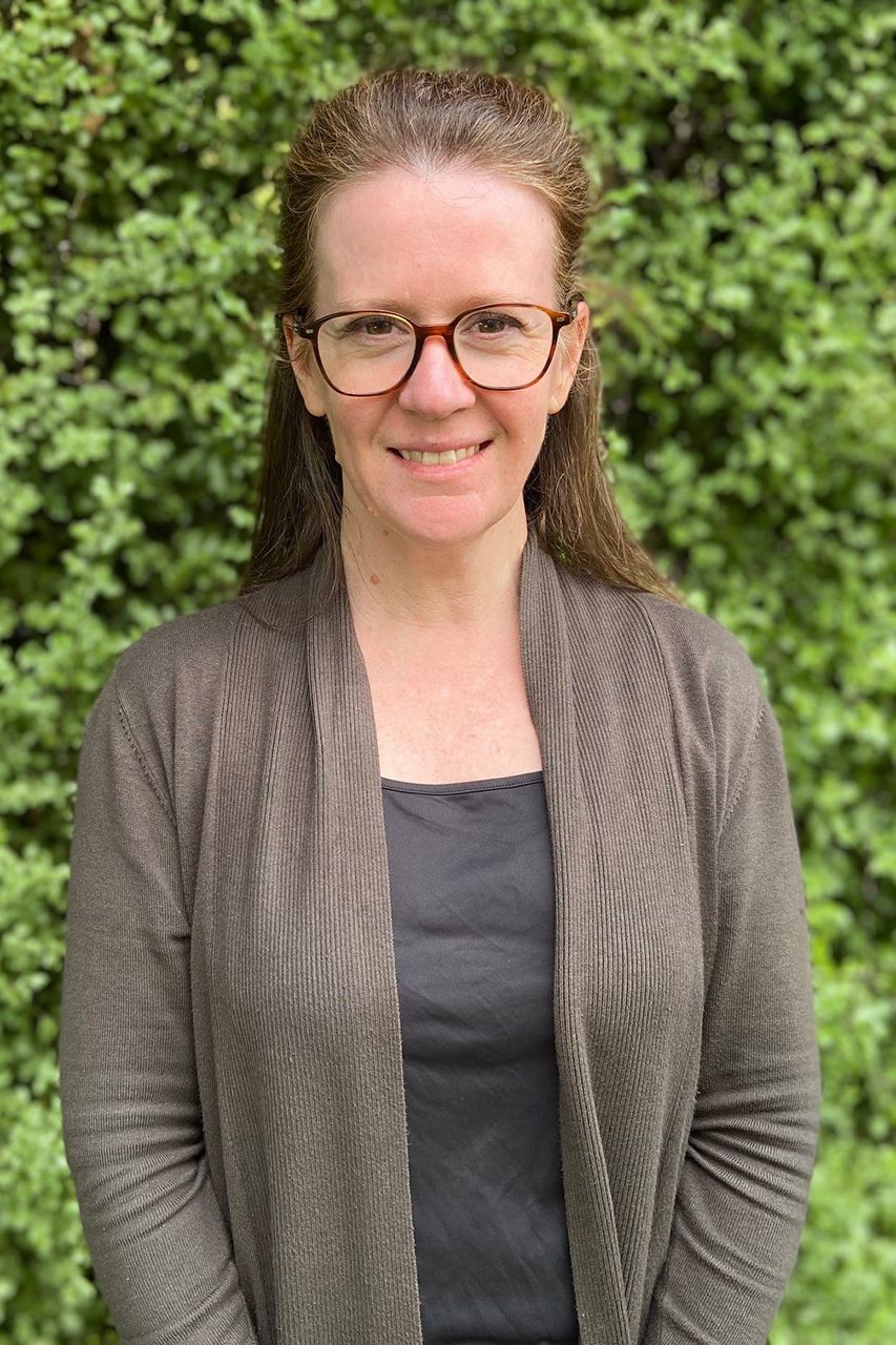 Narelle has long brown hair, wears glasses and has a brown cardigan on.