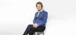 An older woman dressed in a blue jacket and black pants sits in a relaxed pose on a stool