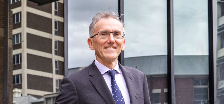 A man dressed in a suit stands in front of buildings in the city and smiles at the camera