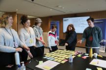 A group of six people of varying ages stand around a table covered in sticky notes and point. 