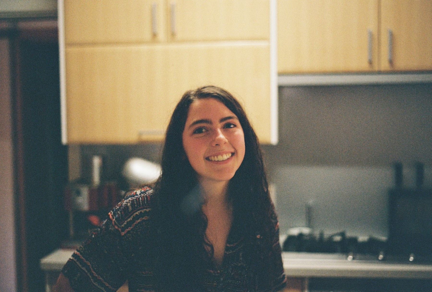 A young female student with brown hair, standing and smiling in a kitchen. 