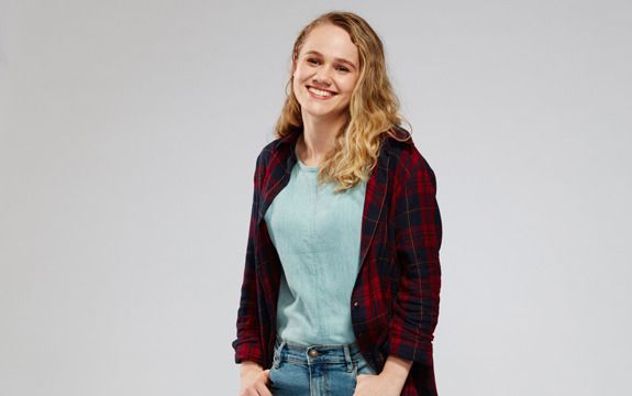 A smiling female student with wavy blonde hair and her hands in her jean pockets.