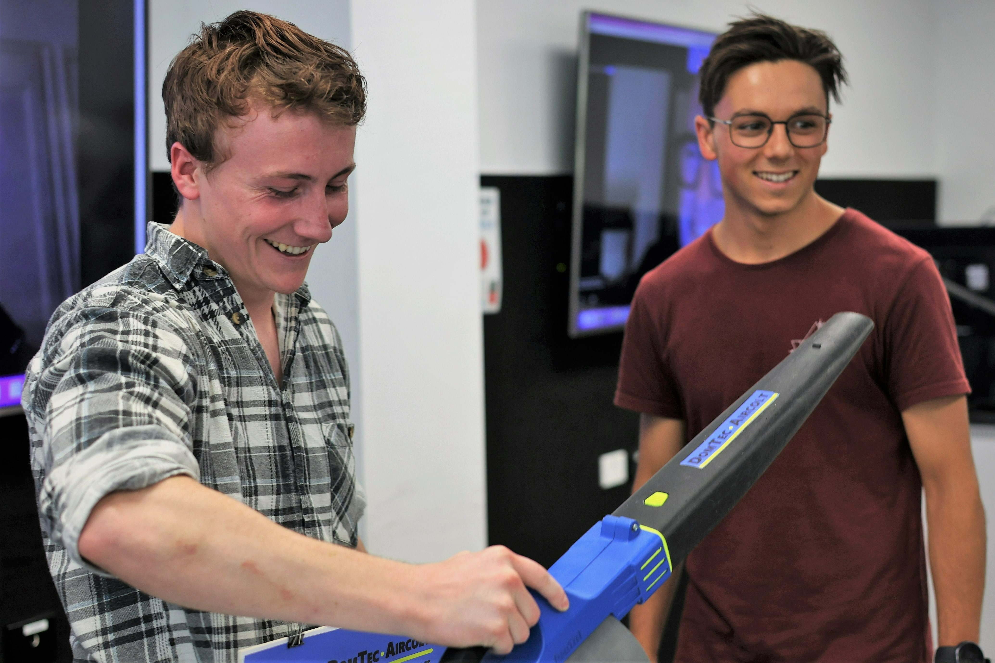 A student in a plaid shirt holds a colorful leaf blower while a second students stands off to the side smiling