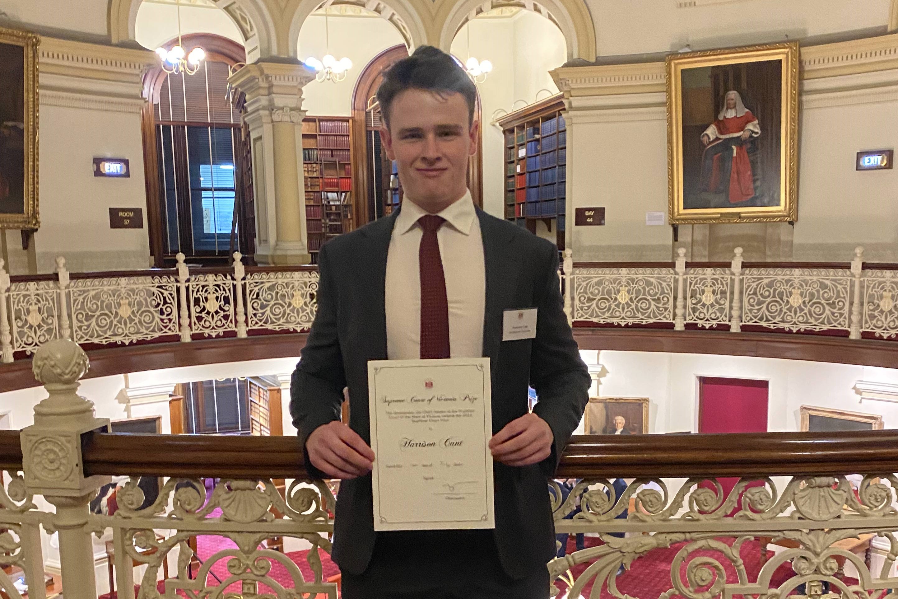 A young man in a suit and tie holds up a certificate in front of an ornate courtroom balcony 