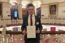 A young man in a suit and tie holds up a certificate in front of an ornate courtroom balcony 