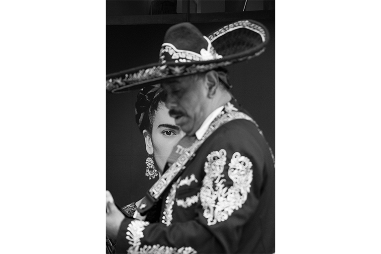 Black-and-white photo of a mariachi musician with a person partially visible behind them.