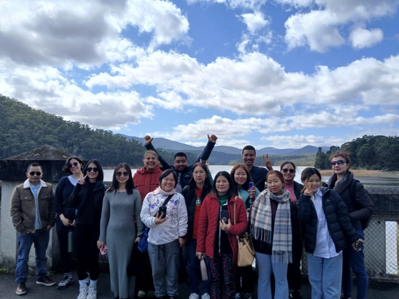 A group of Migrant English Students smile in front of a reservoir of water. In the distance there are tree covered mountains.