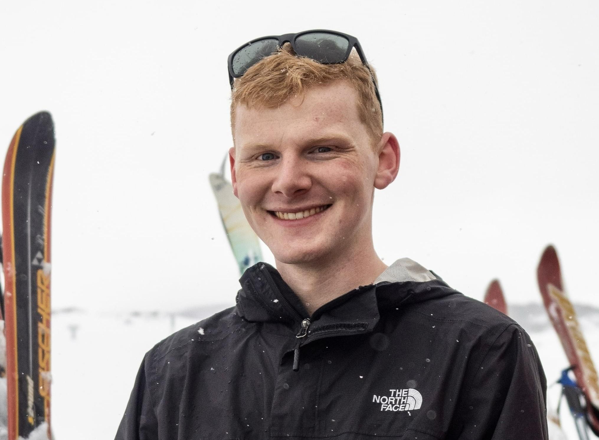 A red-haired student in a black snow jacket, smiling against a backdrop of skis and snow.