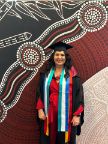 Woman in graduation regalia standing in front of a dot painting mural, smiling.