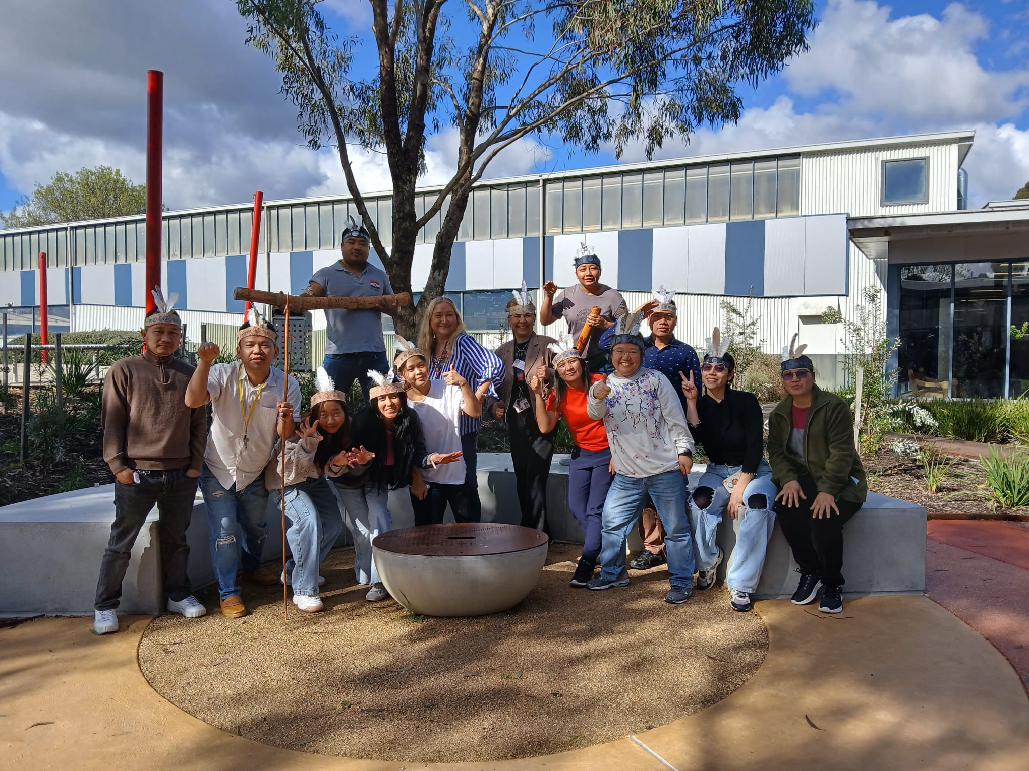 A group of students pose joyfully wearing ceremonial headbands and armbands. They are standing in the Indigenous Learning Circle which has earth coloured circles on the ground and a fire pit.