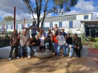 A group of students pose joyfully wearing ceremonial headbands and armbands. They are standing in the Indigenous Learning Circle which has earth coloured circles on the ground and a fire pit.