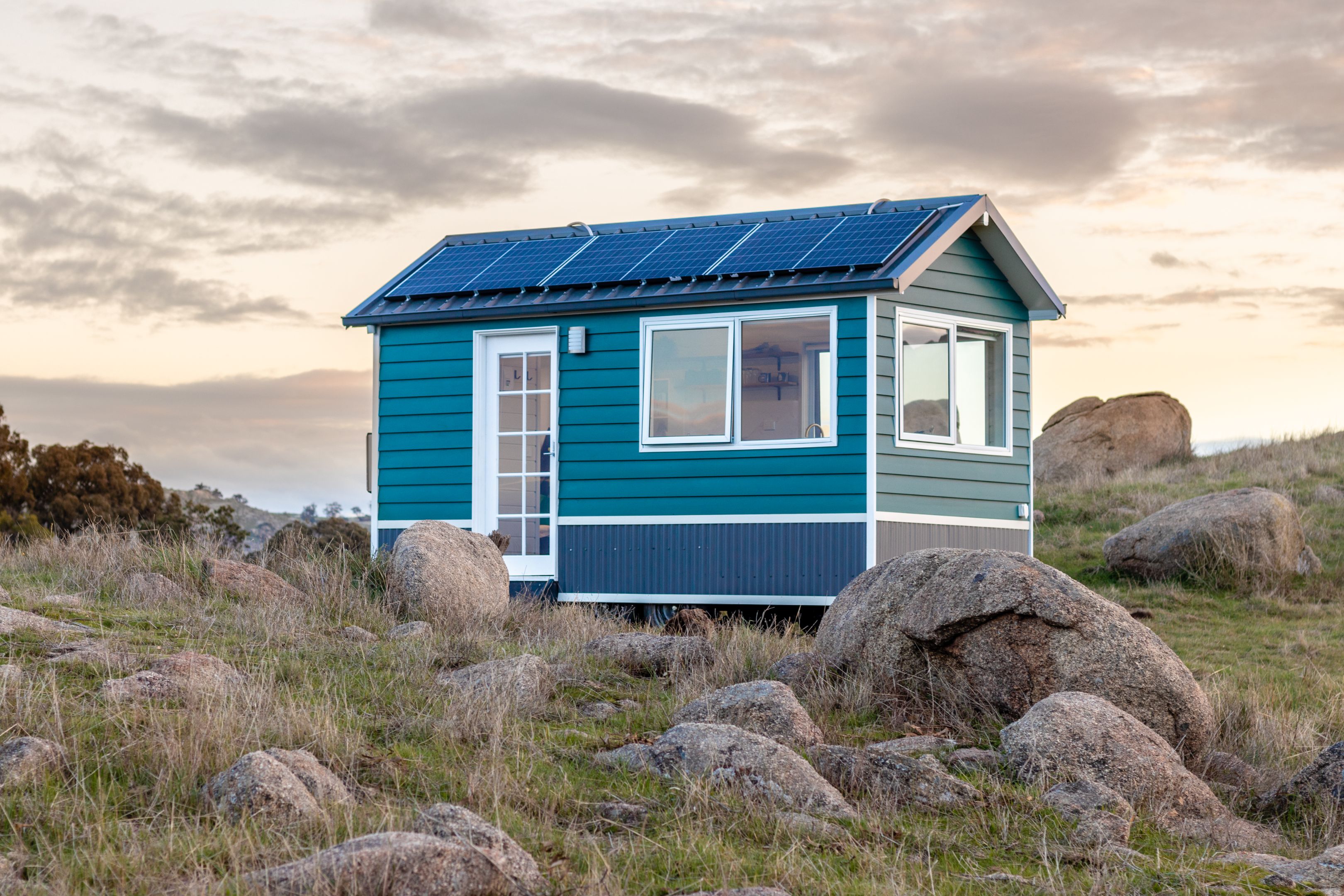 A tiny blue cottage is nested among large rocks, green grass and a rich sunset