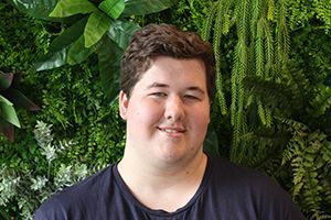 A man wearing a navy t-shirt with brown hair smiles to camera surrounded by green foliage