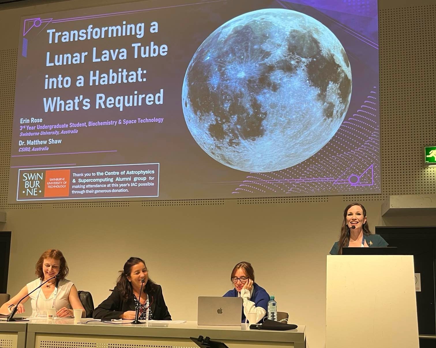 Erin Rose presenting at a podium with the projector behind her. There are three other ladies sitting beside her while she presents