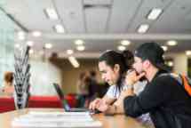 Two students sitting at a desk, looking at a laptop together.