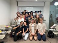 A group of young people pose together in front of a white board in a classroom. Some of them are wearing graduation caps and holding diplomas