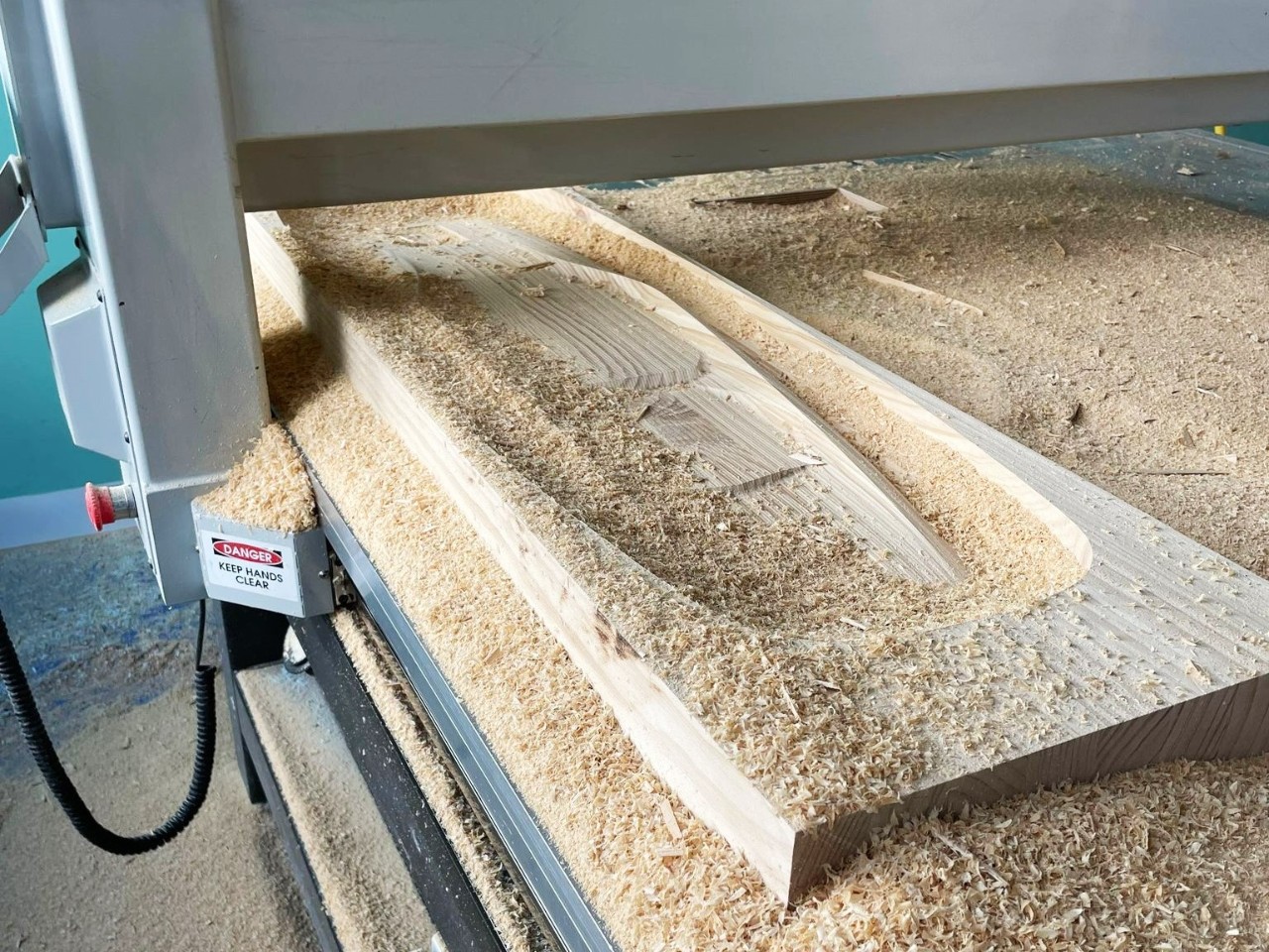 A rectangular wooden piece sits in an industrial machine with a shape cut out of it, surrounded by sawdust