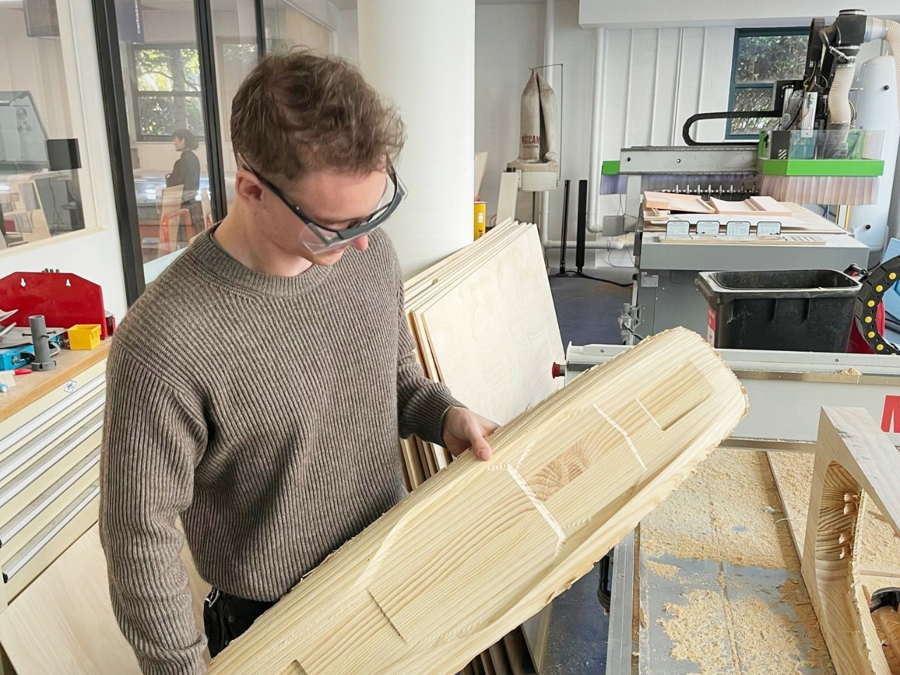 A student in waring safety glasses holds a roughly wooden shape, similar to a skateboard 