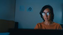 Woman in dark room, looking at computer screen. You can see the reflection of data from the screen on her glasses.