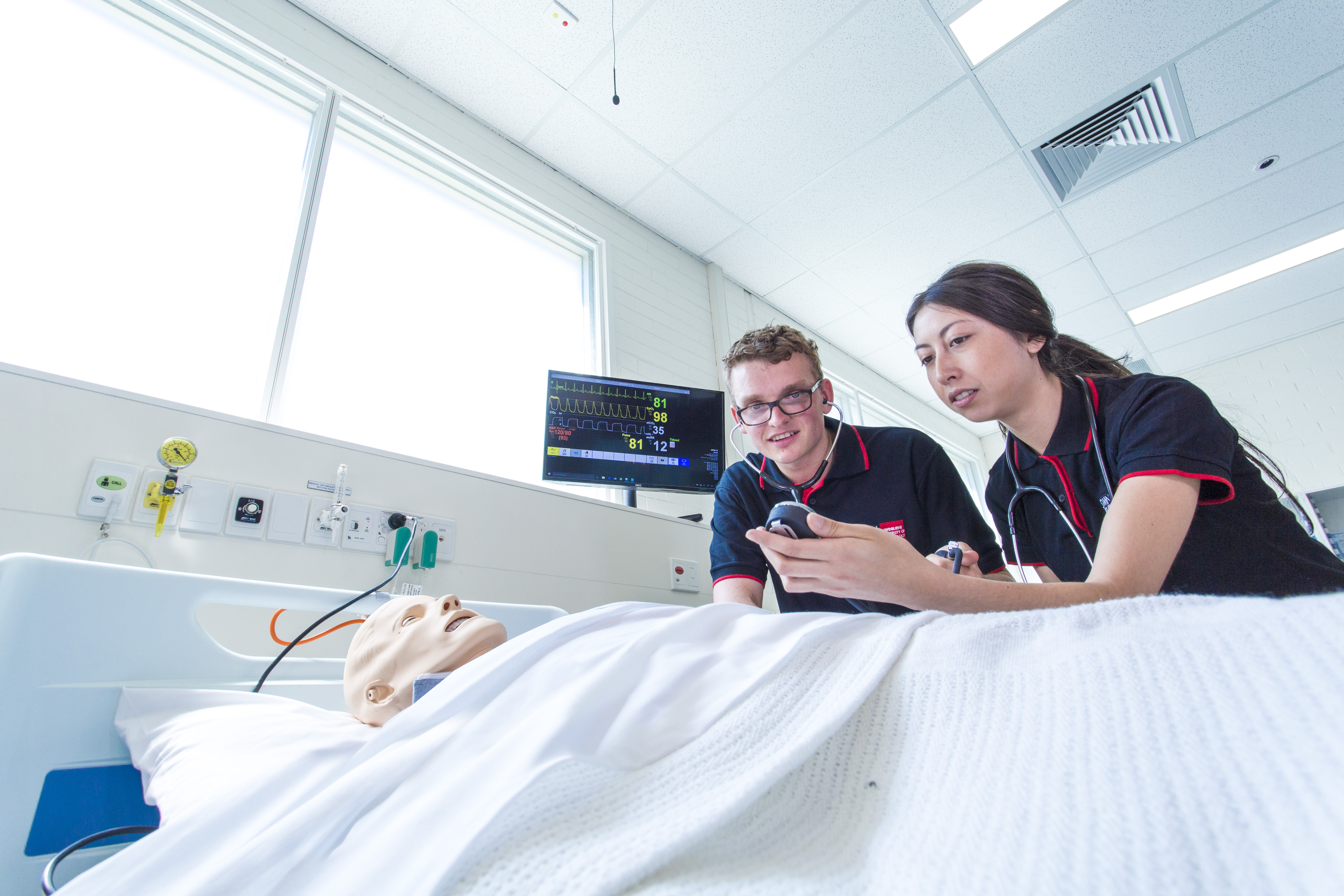 Two nursing students look at medical device beside a dummy patient lying in hospital bed