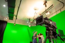 A large black camera with students and a green screen in the background