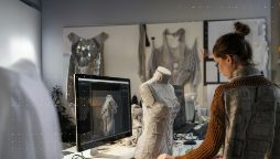 Woman standing on the right hand side of the image with her back to camera, working on a mannequin in a textiles/design room. 